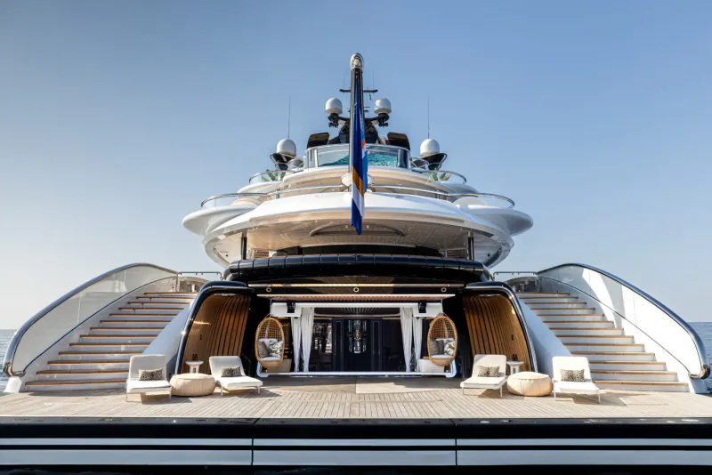 Front view of luxury yacht Kismet with white hull, blue French flag, sun loungers, and stairs on expansive deck against blue sky