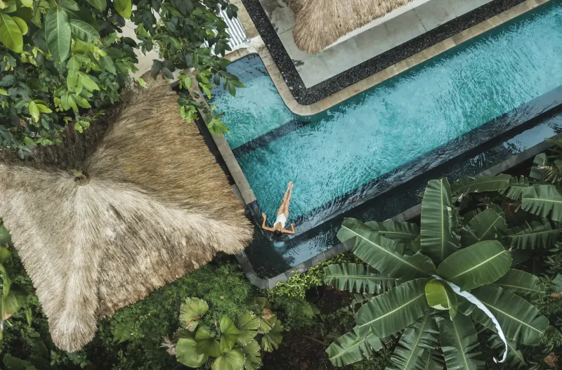 Aerial view of person floating in infinity pool at Bali luxury villa with thatched roofs and lush tropical plants