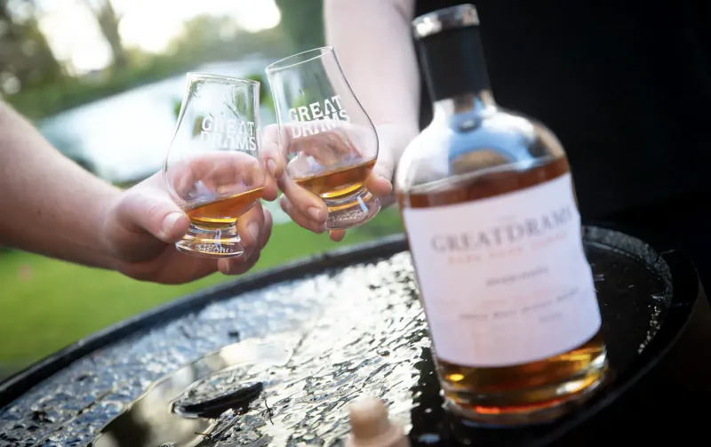 Two hands clinking GreatDrams whisky glasses beside a GreatDrams single malt bottle on a tray outdoors.