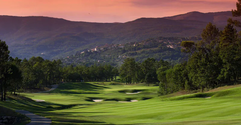 Sunset view of lush green golf course fairway and bunkers amid pine trees and mountains at Terre Blanche resort, Provence.