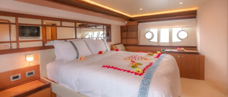 Luxury yacht bedroom with white bed adorned in red hibiscus petals and blue ribbon, porthole windows, warm lighting.