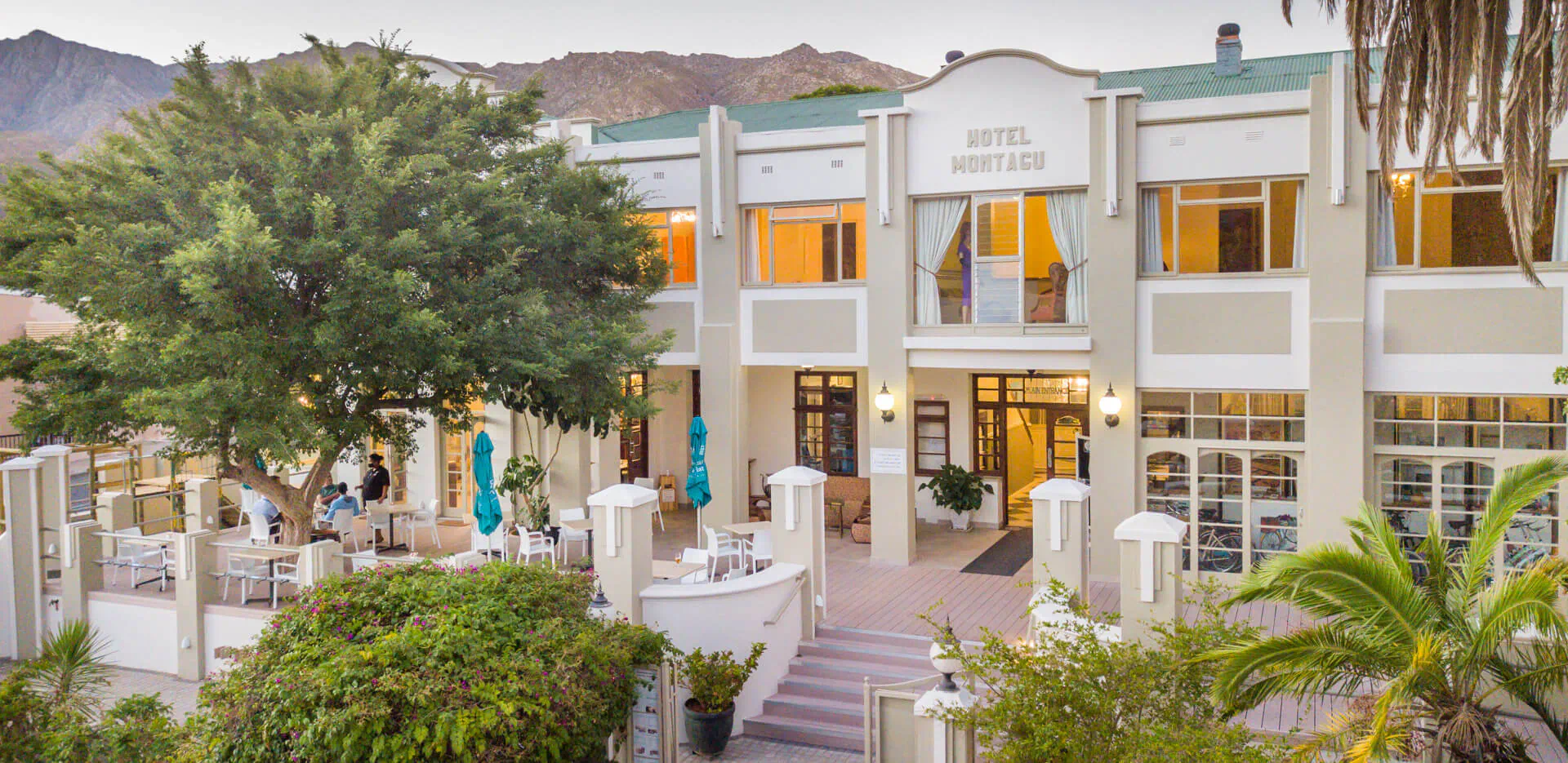 Montagu Country Hotel facade with outdoor seating, palm trees, and mountains in background.