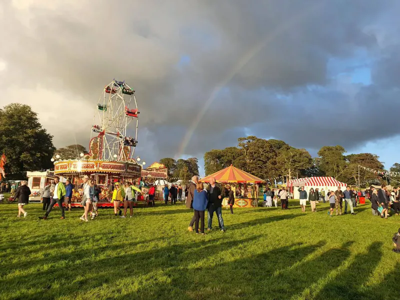 Crowded outdoor festival with Ferris wheel, colorful stalls, people on grassy field under rainbow and cloudy sky