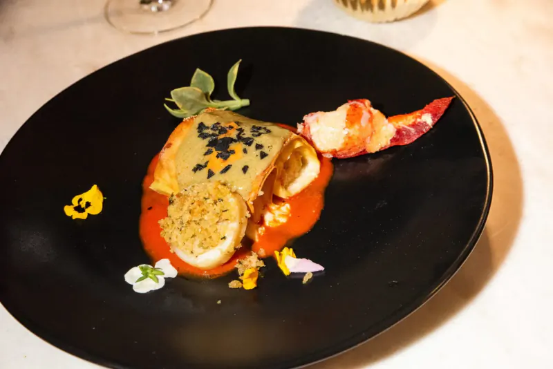 Black plate with sesame ravioli, lobster claw, red sauce, edible flowers, and herbs beside wine glass