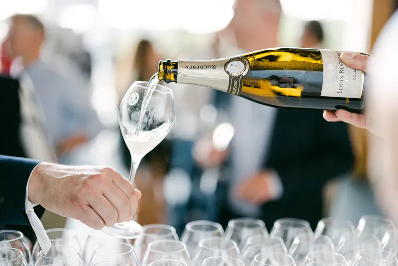 Hand pouring champagne from Moët & Chandon bottle into wine glass amid row of glasses at summer event