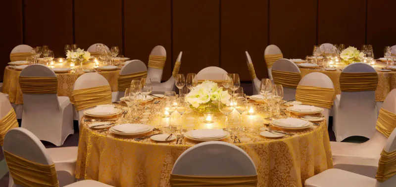 Elegant round dining tables with gold sashes, white orchids, glassware, and candles in luxury ballroom setting