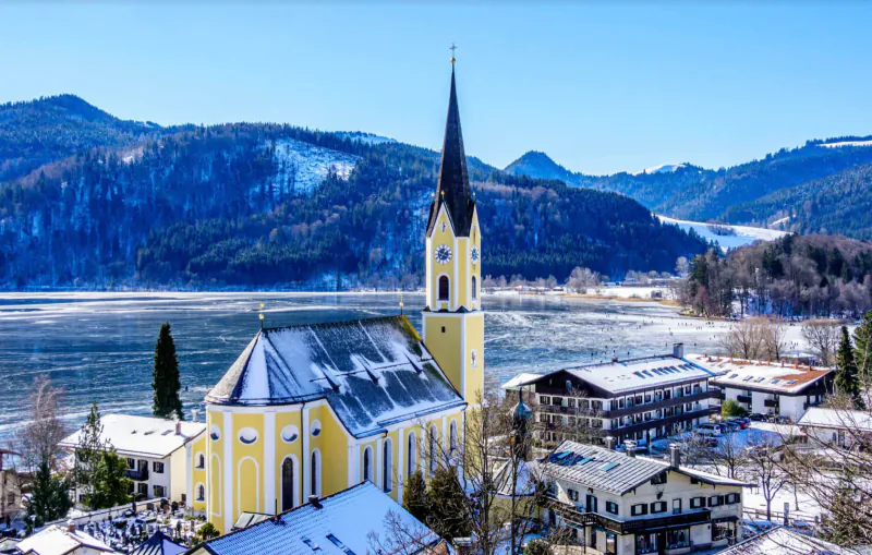 Yellow church with tall spire in snowy Schliersee village, Karma Bavaria, surrounded by Alps and lake in winter.