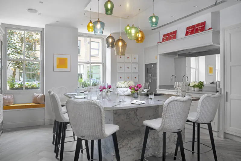 Elegant modern kitchen with white woven chairs around gray marble table set for dining, colorful pendant lights, flowers, large windows.