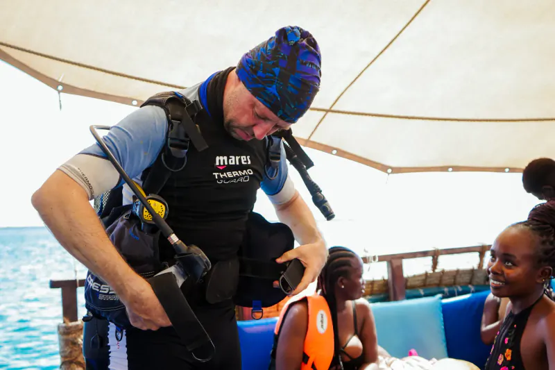 Diver in Mares gear and beanie adjusts equipment on dhow boat in Kisite Marine Park, smiling kids in life vests nearby, ocean view.