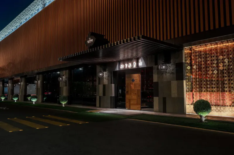Night view of Myazú restaurant entrance with lit 'Myazú' sign, wooden door, ornate wall hangings, and landscaped exterior.