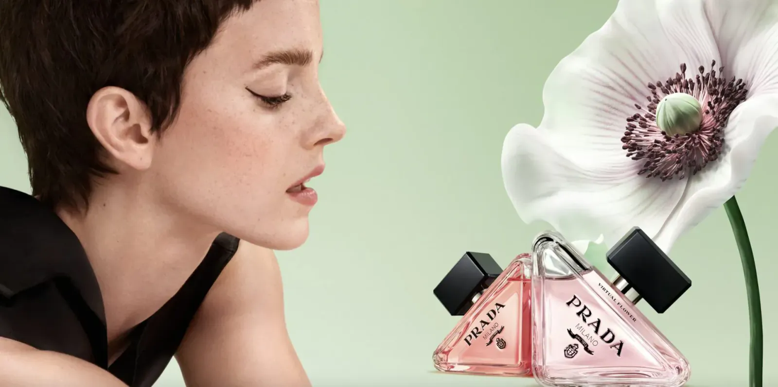 Woman with short hair sniffs large white poppy flower beside two pink Prada perfume bottles on green background