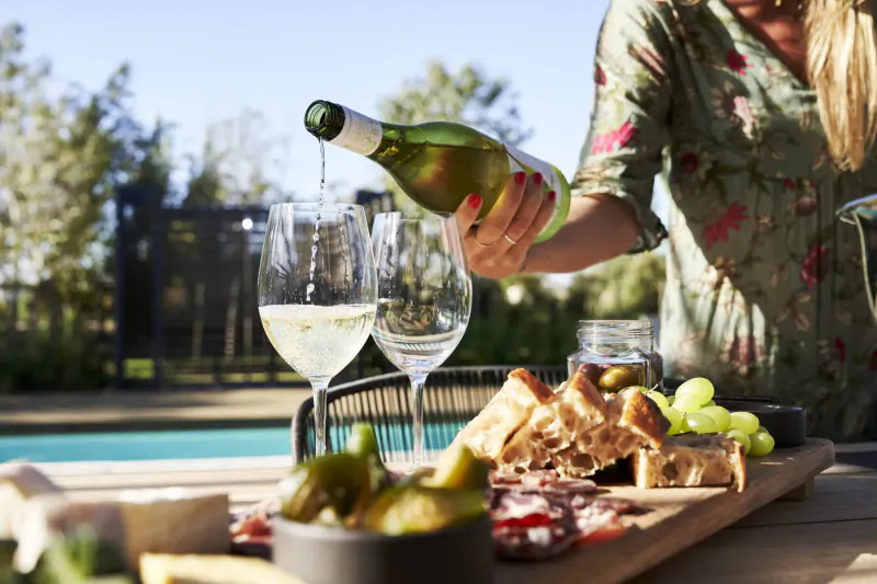 Woman pouring white wine from green bottle into two glasses beside charcuterie board at poolside, Vergenoegd Löw Estate.