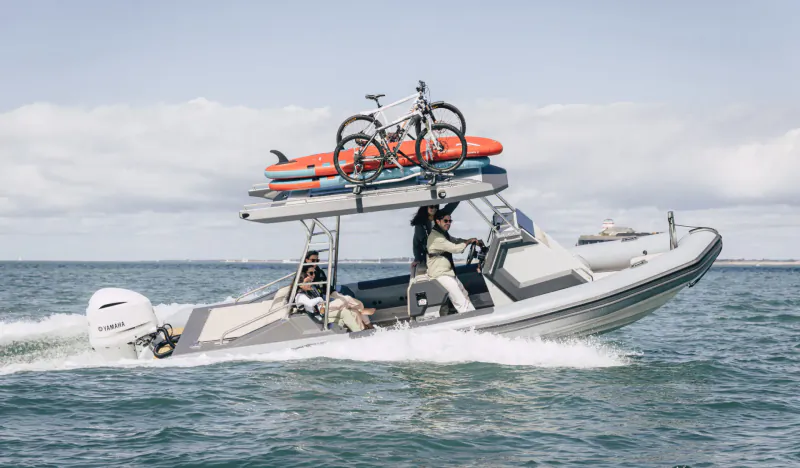 White RIB speedboat speeding on sea with orange surfboards and bikes on roof rack, two crew aboard.