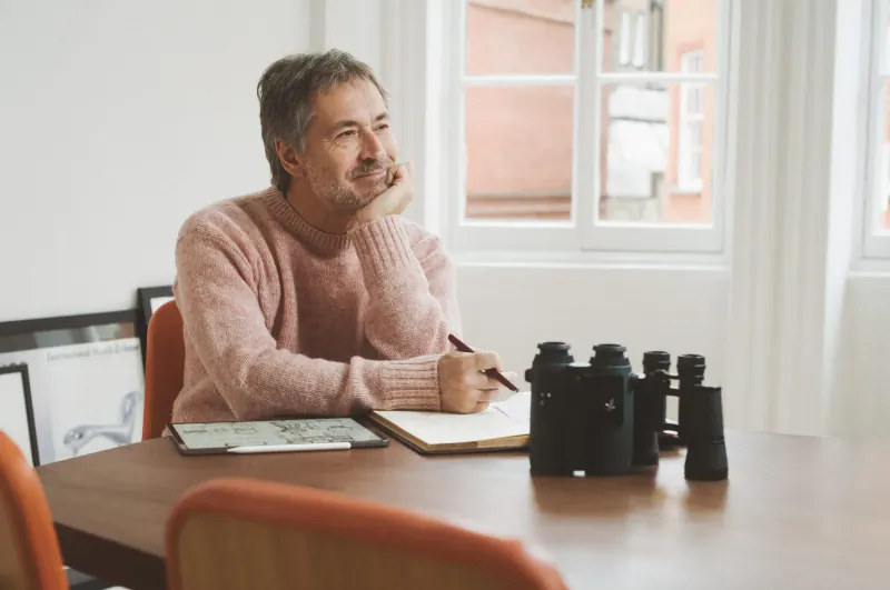 Man with gray hair in pink sweater thoughtfully writing in notebook at desk with smart binoculars and window view