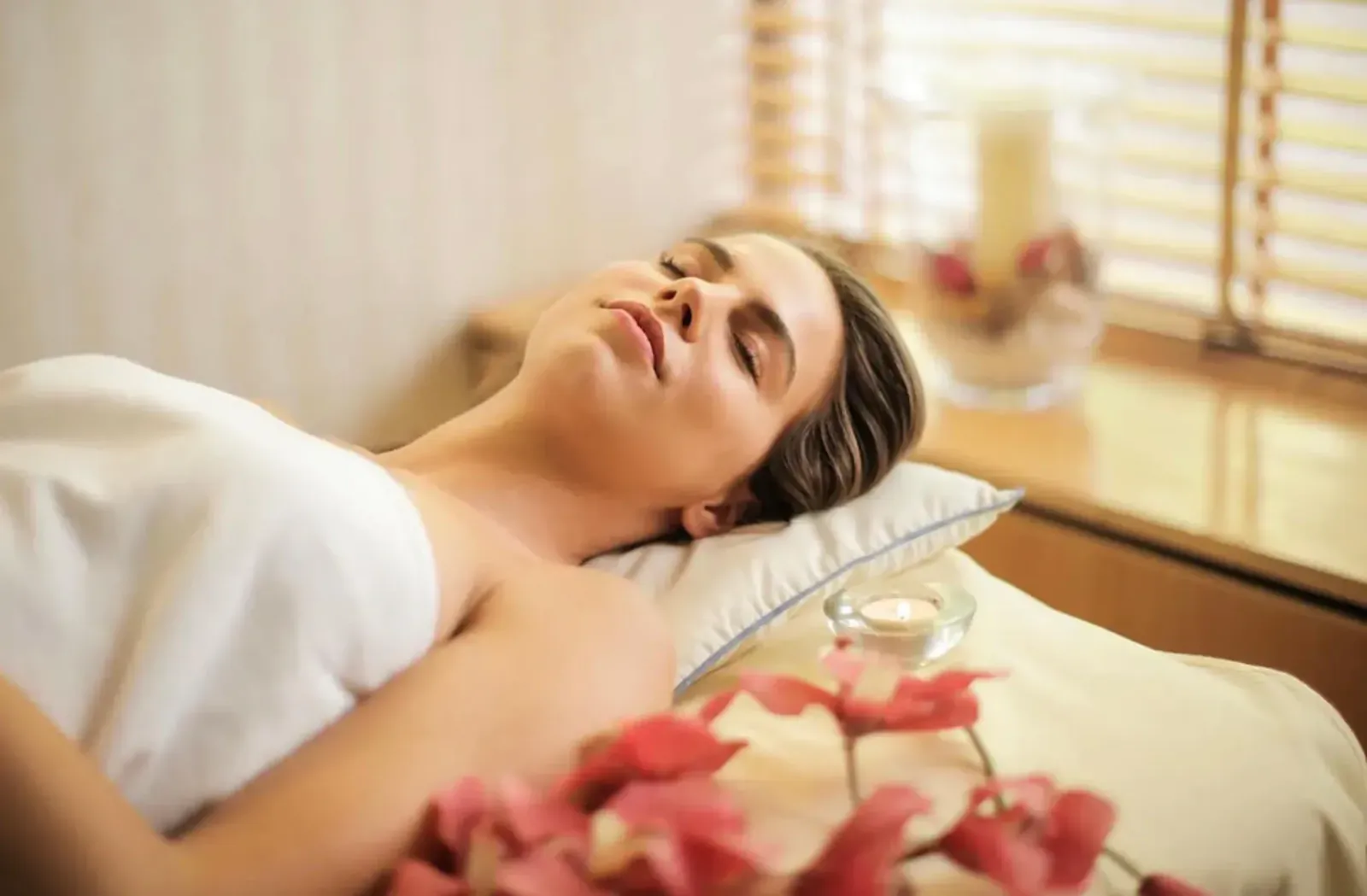 Serene woman in white towel lies relaxed on spa bed with pink orchids, lit by window.