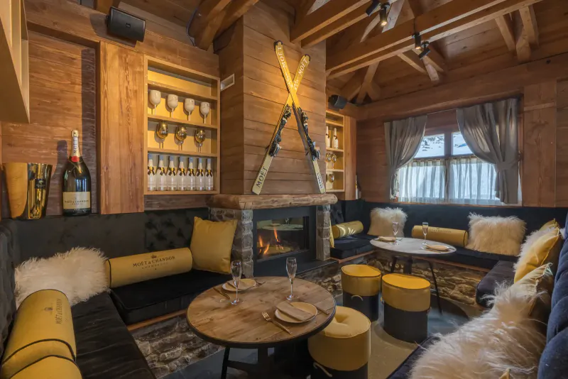 Cozy wooden ski chalet lounge with crossed skis on wall, champagne bottles, lit fireplace, plush seating, and small tables.
