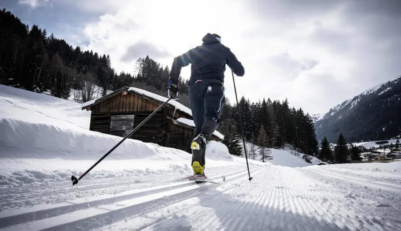 Cross-country skier in black gear speeds along snowy track past wooden cabin in snowy St. Anton mountains.