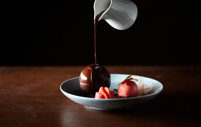 White pitcher pouring chocolate sauce over chocolate sphere dessert with strawberry on plate