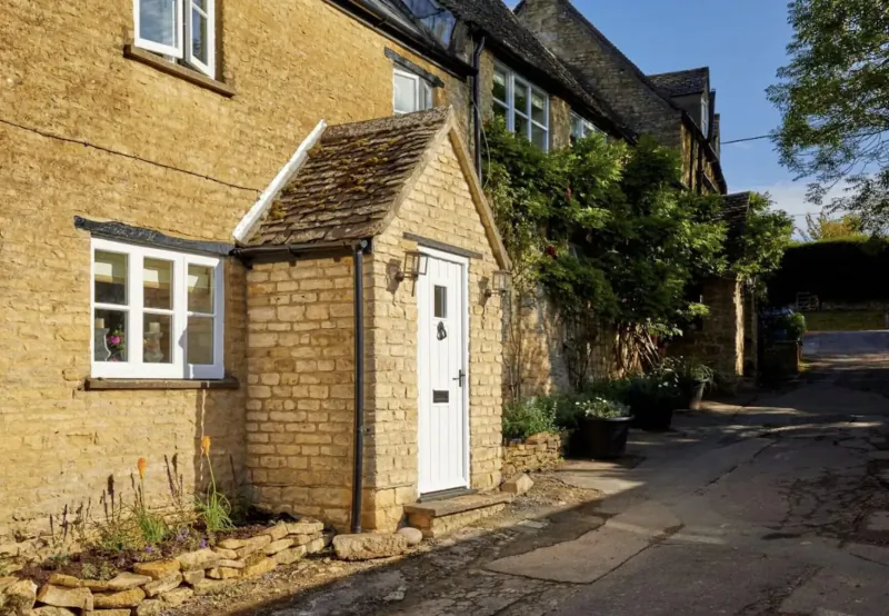 White door on stone porch of Cotswolds cottage, ivy-covered walls, plants, narrow lane, clear sky.