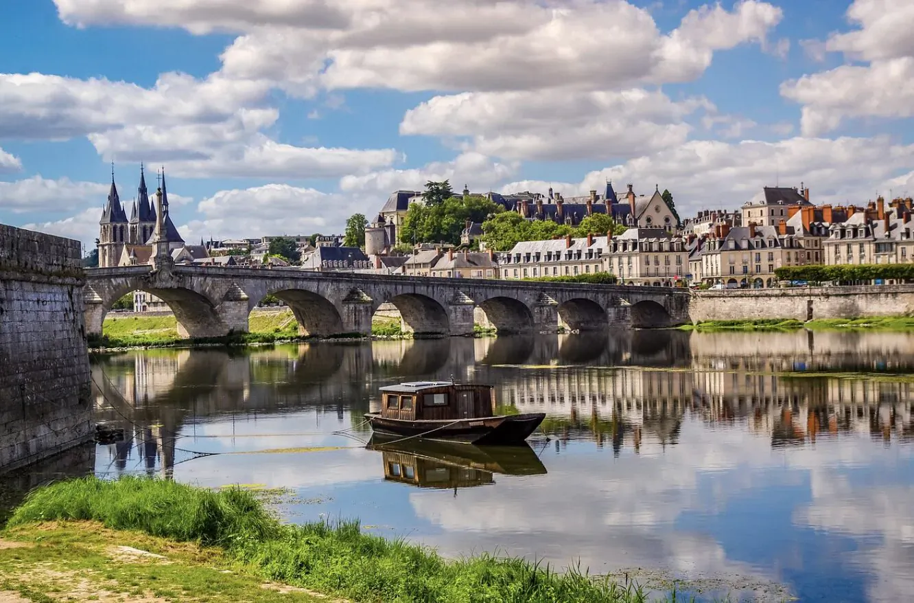 Saint-Bénézet Bridge with towers spanning calm river, small boat on water, historic buildings, blue sky and clouds.