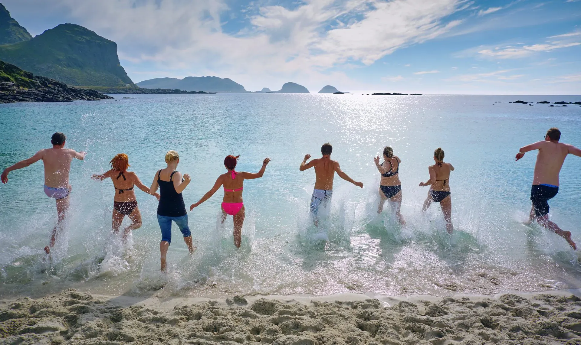 Group of diverse young adults in swimsuits running joyfully into turquoise ocean waves at sunny beach with mountains.