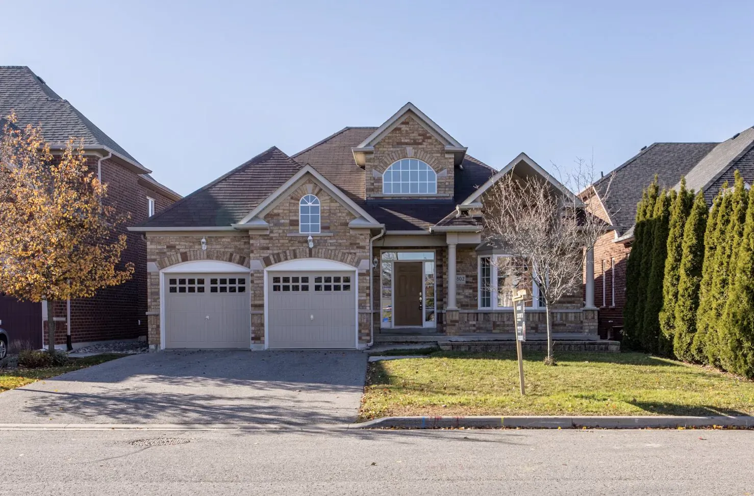 Brick two-car garage house with stone accents, driveway, lawn, hedges, autumn trees on sunny day