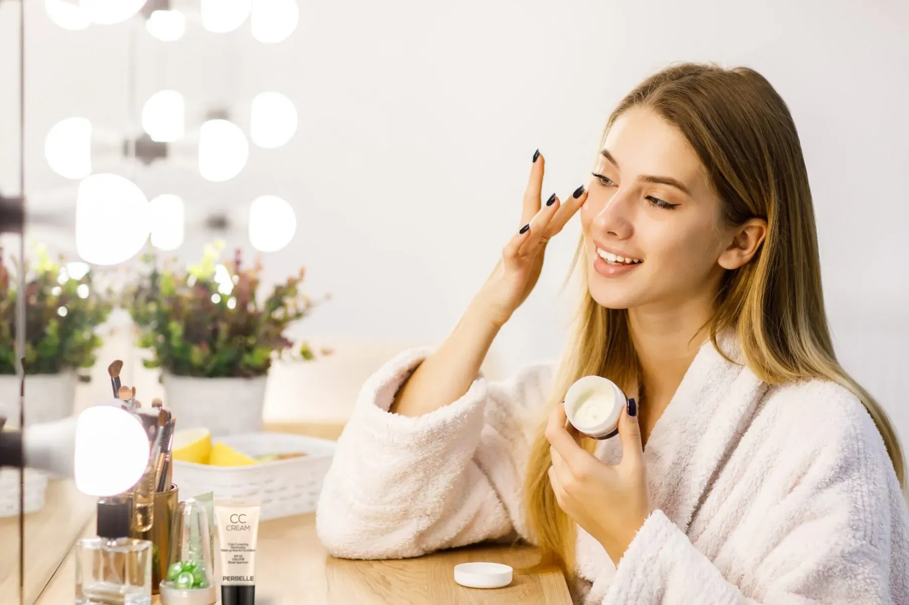 Smiling woman in white robe applies white cream from jar to cheek at vanity mirror with lights and skincare products