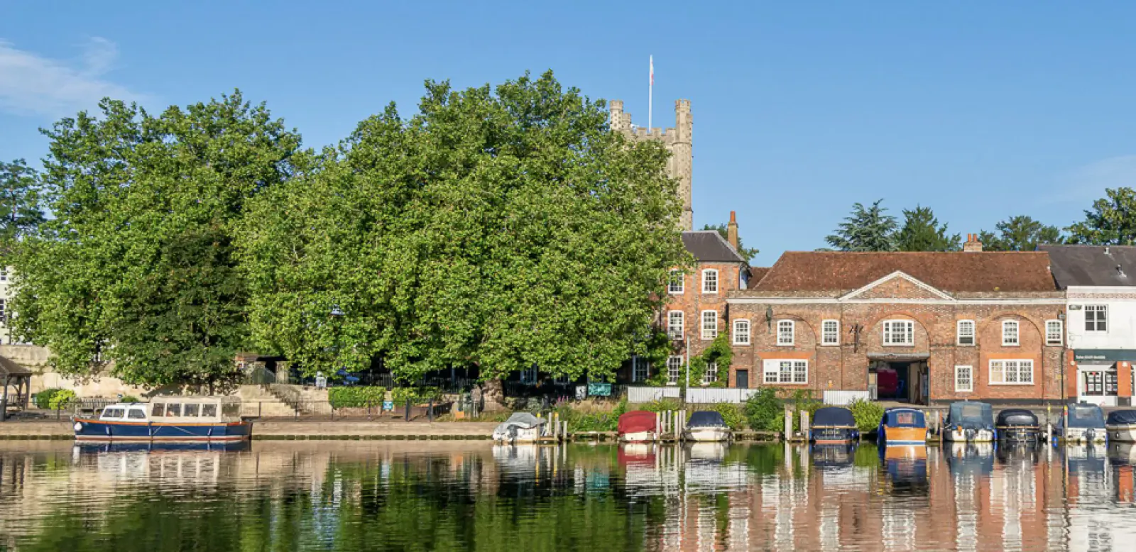 Riverside scene at The Relais Henley: colorful houseboats moored by brick buildings, church tower, and trees under blue sky, with reflections.