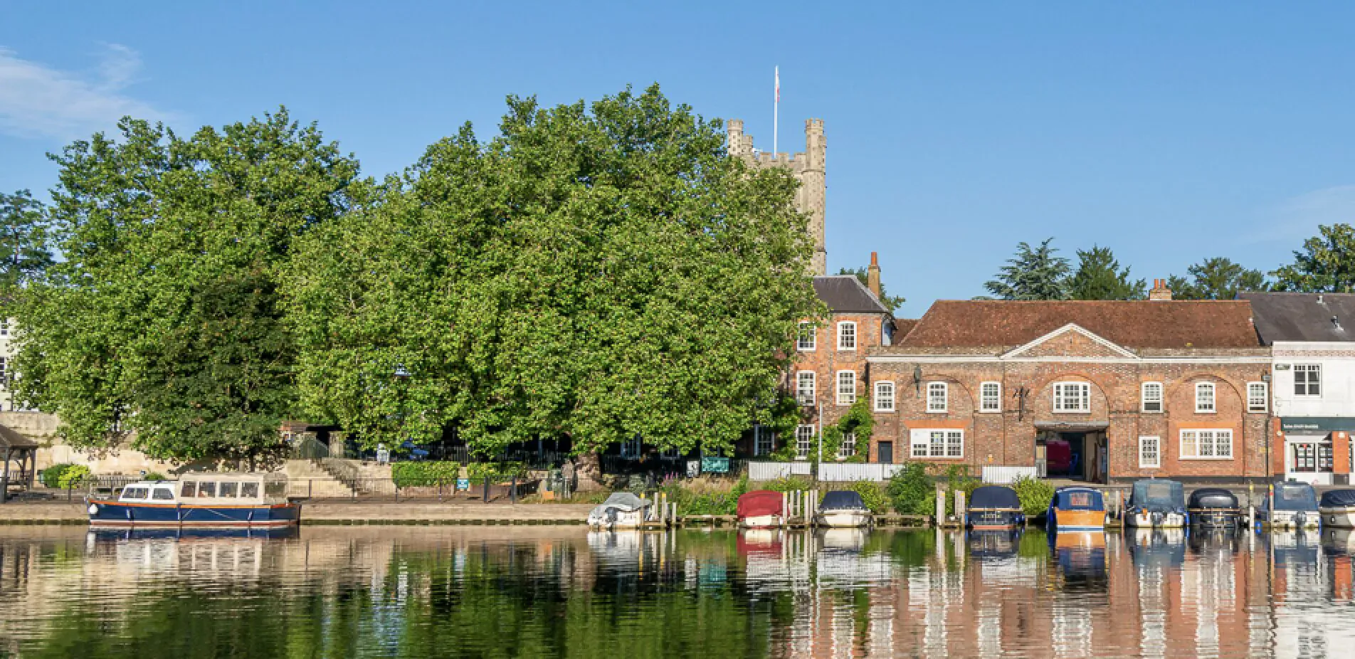 Riverside scene at The Relais Henley: colorful houseboats moored by brick buildings, church tower, and trees under blue sky, with reflections.