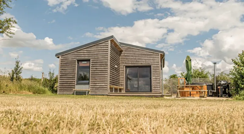Wooden luxury lodge with hot tub beside it in grassy field, surrounded by trees under partly cloudy sky.