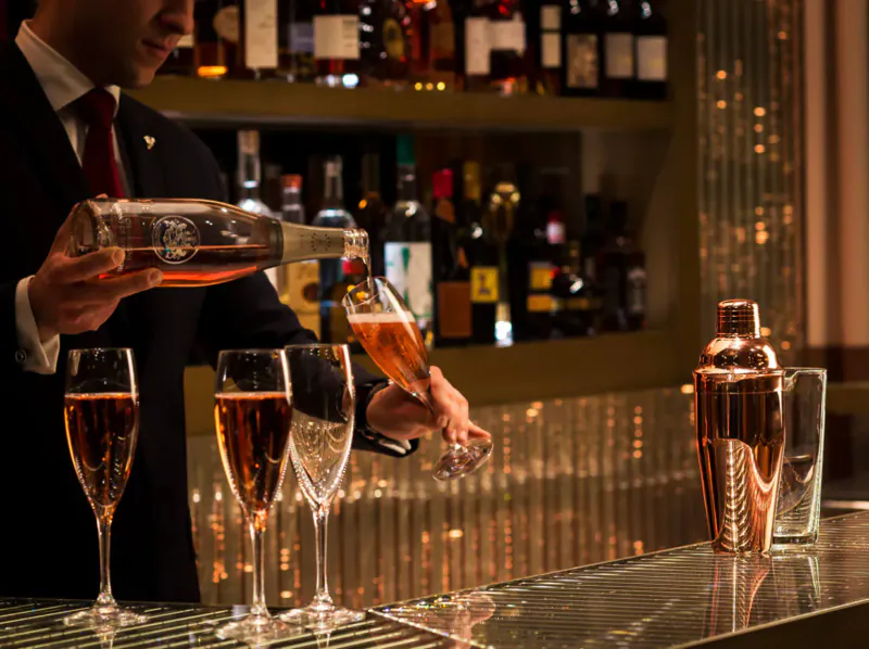 Bartender in suit pouring amber liquor from bottle into champagne flutes at upscale bar with backlit shelves of bottles