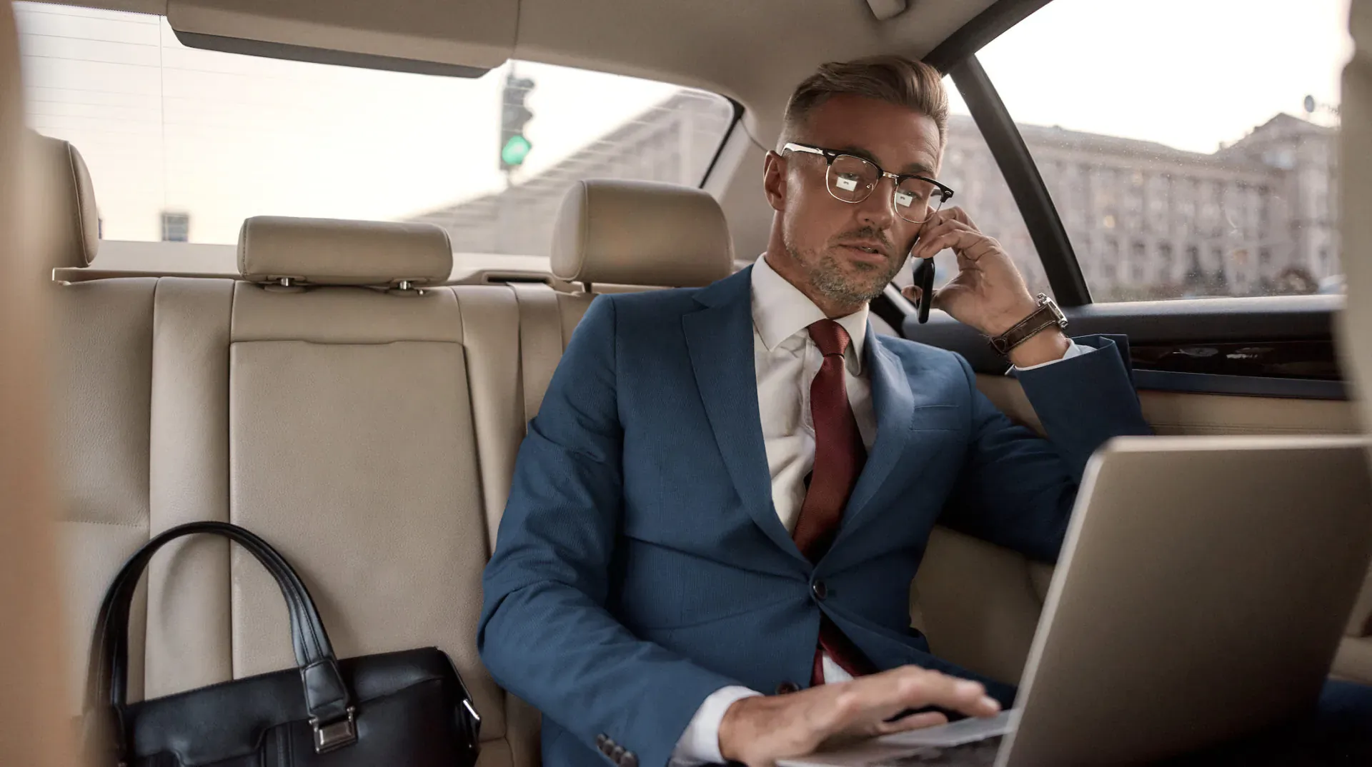 Businessman in suit with glasses talks on phone while typing on laptop in luxury car backseat, city view outside.
