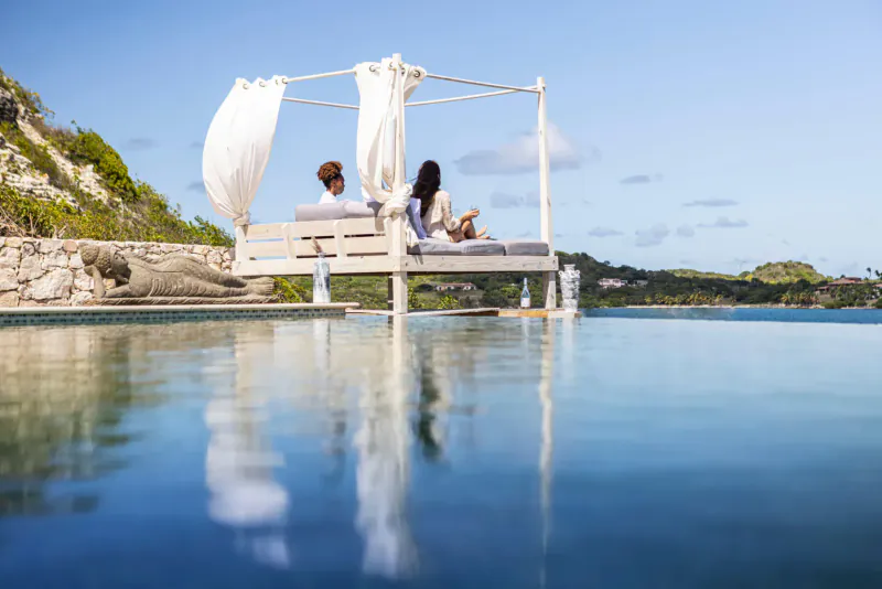 Couple relaxing on a white canopy daybed over infinity pool at Rokuni, Antigua, with ocean and cliffs.