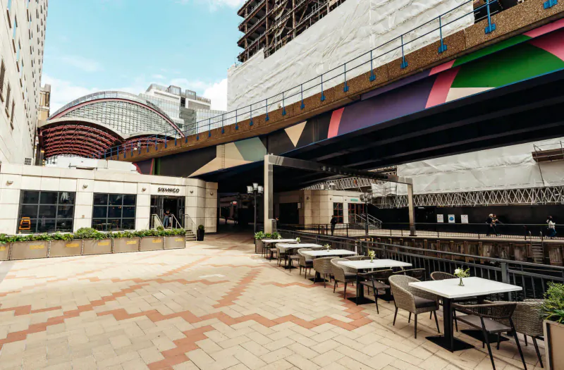Outdoor seating area at Six by Nico Canary Wharf with colorful geometric pedestrian bridge and modern buildings under blue sky