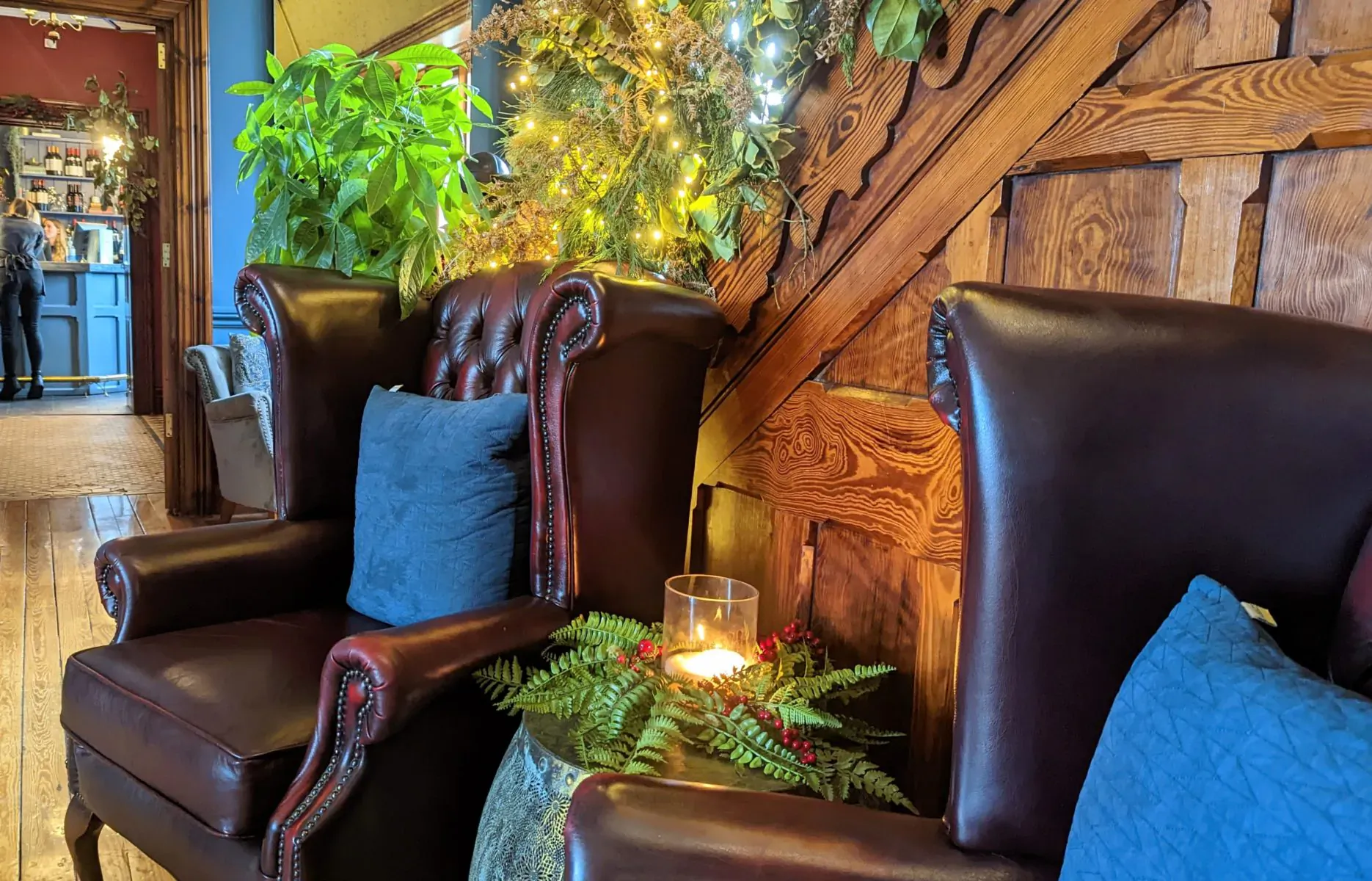 Two red leather armchairs with blue cushions by a lit candle and ferns in a cozy wooden-paneled room with Christmas tree.