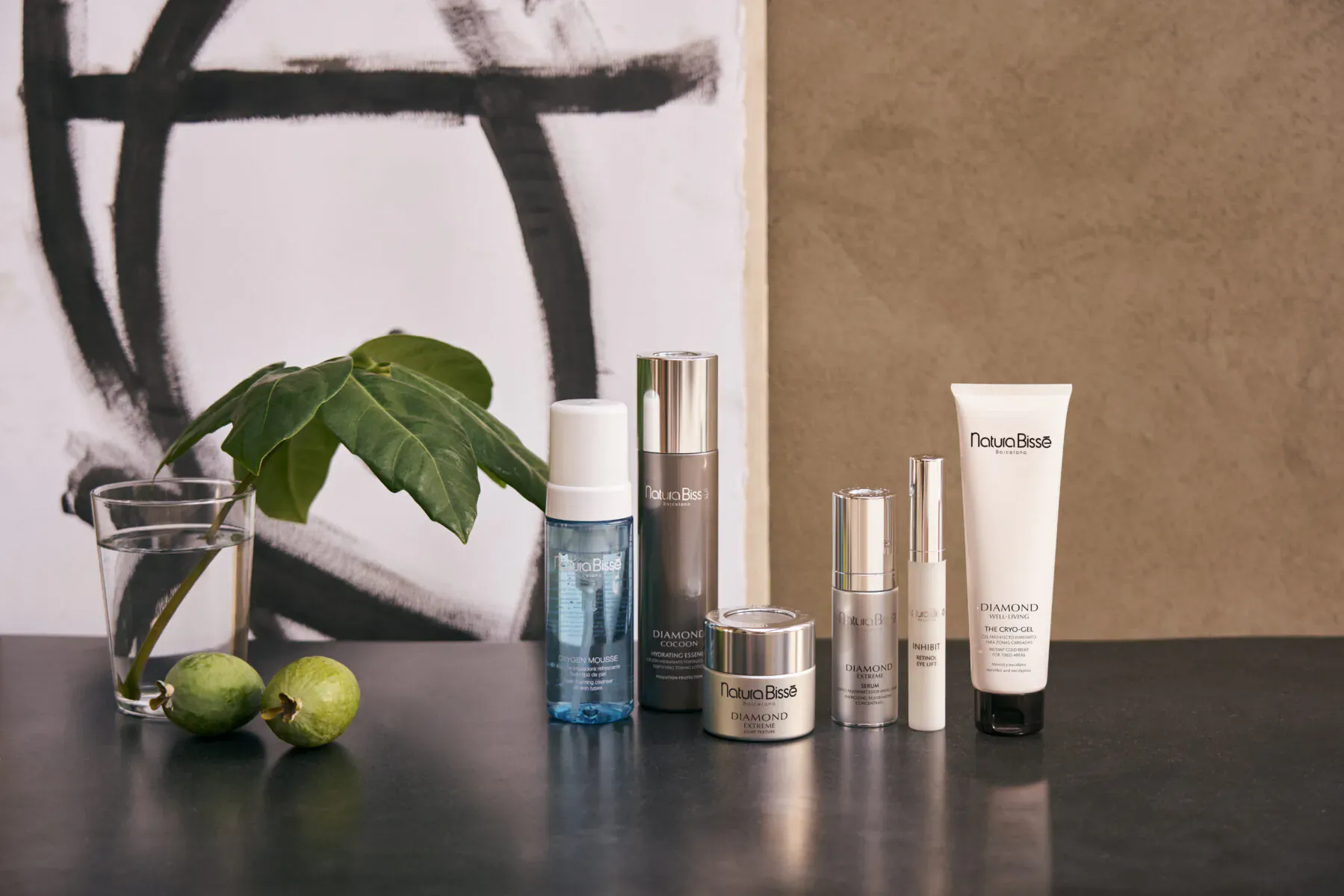 Natura Bissé skincare products on black table with green leaves, limes, water glass, and abstract art backdrop