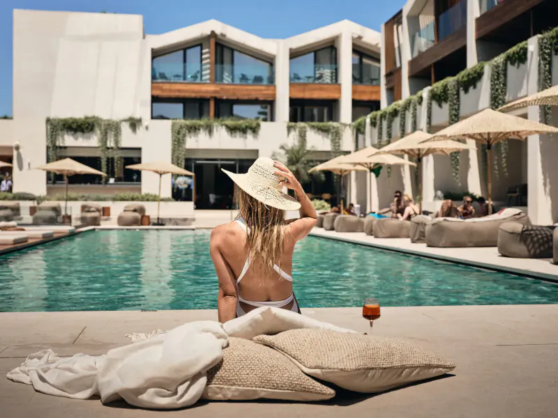 Woman in white bikini and hat sits by Contessina Hotel pool, holding wine glass, luxury resort backdrop.