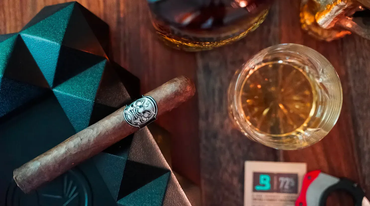 Close-up of lit cigar with skull band, whiskey glass, green geometric box, cigar cutter showing 977, on wooden table