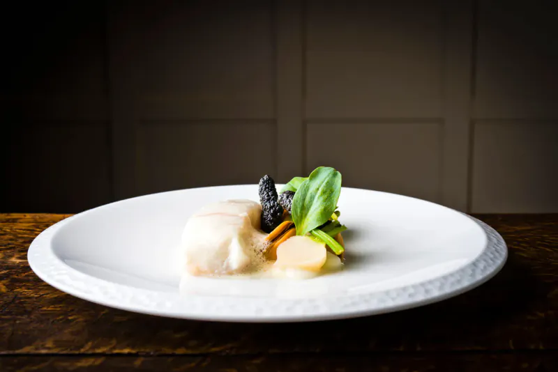 White plate with poached white fish, black truffle, turnips, and greens on wooden table