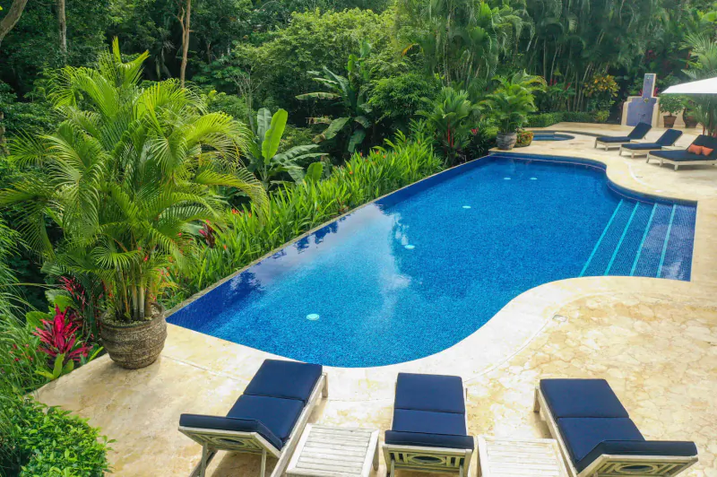 Luxury villa infinity pool surrounded by lush tropical jungle, with blue lounge chairs on beige deck.