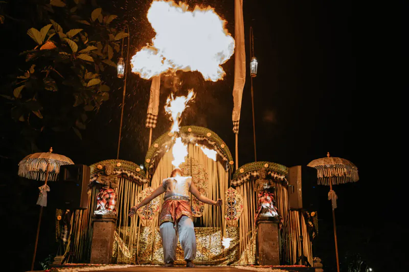 Balinese dancer in traditional attire performs fire breathing on stage at night, framed by ornate golden arches and umbrellas