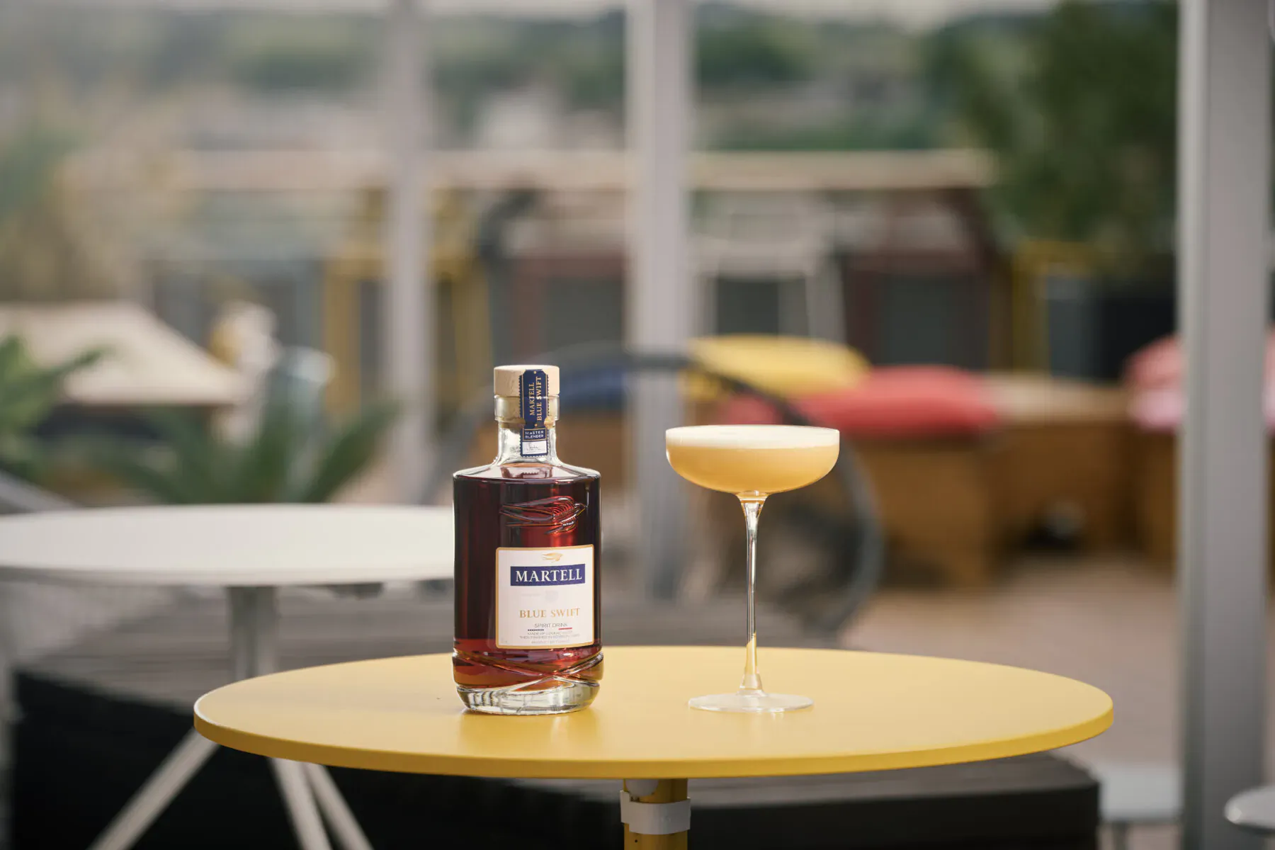Martell cognac bottle and creamy daiquiri cocktail on yellow table at L’Indigo bar with blurred terrace view