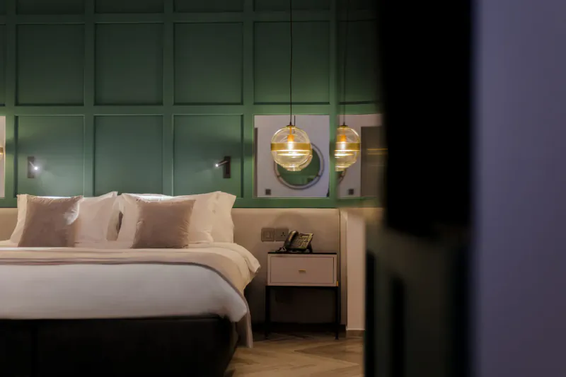 Cozy hotel bedroom with green paneled walls, plush white bed, glowing pendant lights, and nightstand in Embassy Valletta.