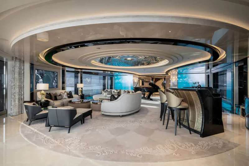 Luxurious yacht interior with curved white sofas, grand piano, bar, and panoramic ocean windows.