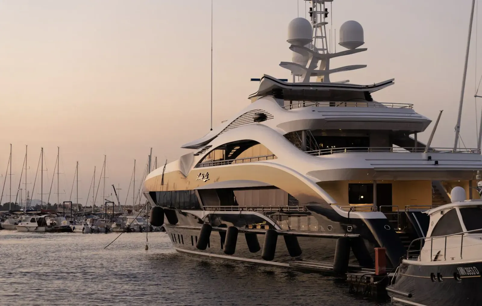 Sleek white and gold luxury yacht docked at marina during sunset, surrounded by sailboats.