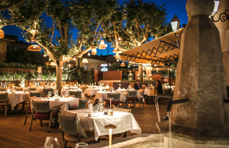 Elegant outdoor dining at Byblos Palace hotel in Saint-Tropez, candlelit tables under trees and lanterns by fountain at dusk