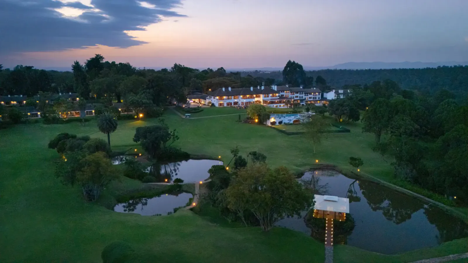 Aerial sunset view of Fairmont Mount Kenya Safari Club with lit chalets, ponds, lawns, and trees.