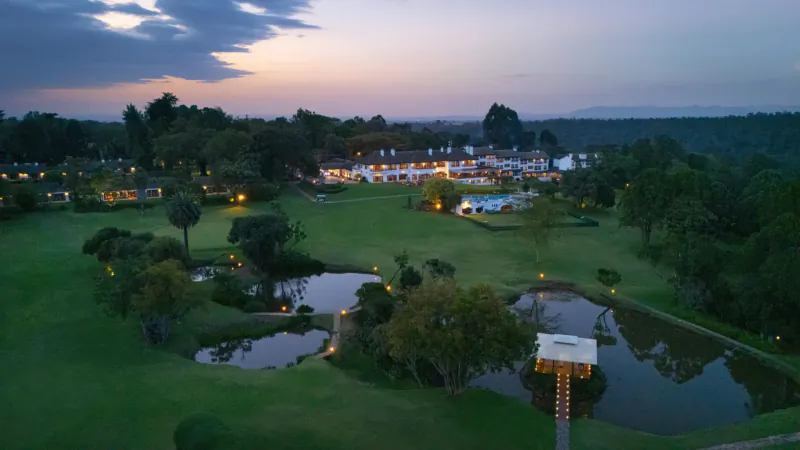 Aerial sunset view of Fairmont Mount Kenya Safari Club with lit chalets, ponds, lawns, and trees.