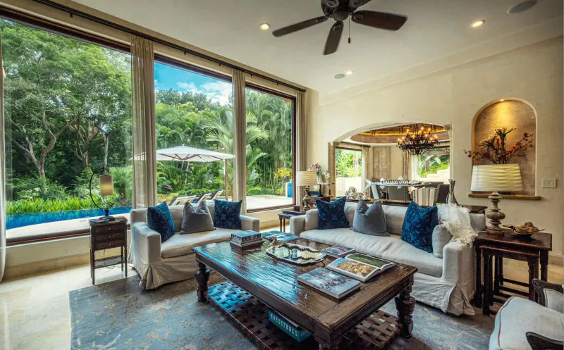 Luxurious living room in private villa with plush blue sofas, wooden coffee table, pool view through large windows.