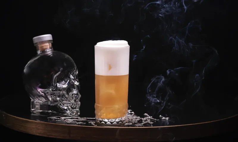Skull-shaped glass bottle beside tall cocktail with foamy white top and amber liquid on dark table with smoke, at rooftop bar.